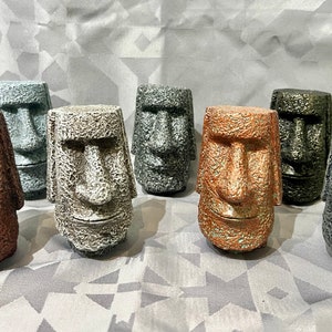 May include: Eight small, textured, stone-like sculptures of Easter Island heads in various shades of grey, copper, and bronze.