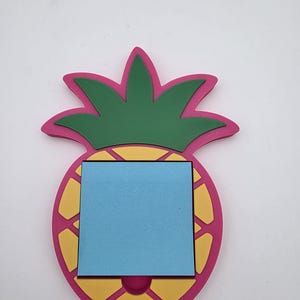 May include: A pineapple-shaped memo board with a pink outline, yellow segments, and green leaves. A blue square notepad is in the center. The memo board is designed to resemble a pineapple.
