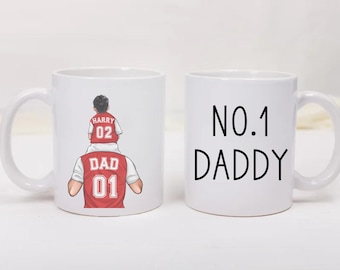 May include: Two white ceramic mugs. One mug features a graphic of a child sitting on a person's shoulders, both wearing red and white jerseys with the text HARRY 02 and DAD 01. The second mug displays the black text NO.1 DADDY in a simple font.