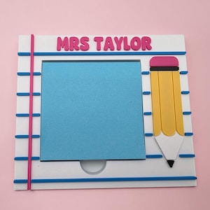 May include: A white notepad-style frame with the text "MRS TAYLOR" in pink letters. It features a blue square for a note, a yellow pencil graphic with a pink eraser, and pink and blue accents. The background is a solid pink.