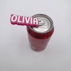 May include: A red Coke Zero can with a white and pink name tag that says "OLIVIA". The can is 330ml and has a silver top.