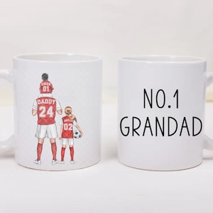 May include: Two white ceramic mugs. One mug features a graphic of a person carrying a child on their shoulders and standing next to another child, all wearing red and white soccer jerseys. The other mug displays the black text NO.1 GRANDAD.