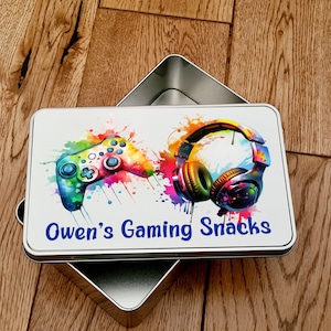 May include: A silver metal tin with a colourful design of a video game controller and headphones. The tin has the text "Owen's Gaming Snacks" printed on it.