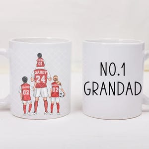 May include: Two white ceramic mugs. One mug features a graphic of a person with three children wearing red and white soccer jerseys, with the text DADDY 24, OWEN 01, JACOB 02, and AMALA 02. The second mug displays the black text NO.1 GRANDAD.