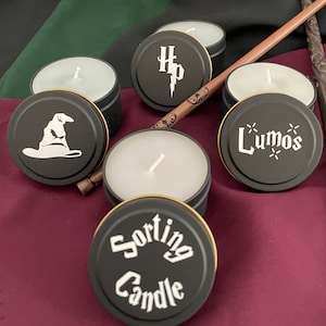 Könnte beinhalten: Eine Sammlung schwarzer Teelicht-Kerzenbehälter mit goldenen Rändern, mit weißen Grafiken eines Sprechenden Huts, dem Harry-Potter-Logo und dem Wort "Lumos". Ein Holzstab ist ebenfalls vorhanden. Die Kerzen befinden sich auf einer weinroten Oberfläche.