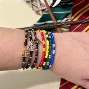 Könnte beinhalten: Eine Sammlung von Perlenarmbändern in verschiedenen Farben, darunter Schwarz, Gold, Rot, Gelb, Grün und Blau. Einige Armbänder haben einen goldfarbenen Deathly Hallows-Anhänger. Die Armbänder werden an einem Handgelenk gezeigt.