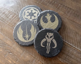 May include: Four black slate coasters with gold-colored Star Wars logos. The logos are the Rebel Alliance symbol, the Imperial symbol, the Mandalorian symbol, and the Jedi symbol.