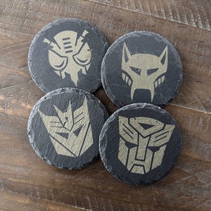 May include: Four black slate coasters with gold-colored Transformers logos. The logos include the Autobot symbol, the Decepticon symbol, Starscream, and Bumblebee.