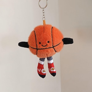 Puede incluir: Un llavero de baloncesto de peluche naranja con una cara sonriente, brazos negros y piernas con zapatillas rojas y blancas. La pelota tiene líneas negras y está unida a un llavero plateado.