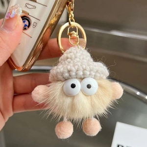 May include: A quirky keychain charm depicting a fluffy, pale-yellow creature with oversized, googly eyes and a knitted white hat. The charm has small pink pom-pom feet and arms, attached to a gold-coloured keyring.
