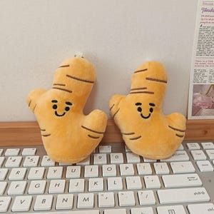 May include: Two plush ginger root toys with smiling faces and brown details. The toys are yellow-orange and sit on a computer keyboard. The background includes a wall and a printed page with text.