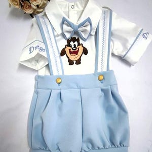 Puede incluir: Una camisa blanca de manga corta con una pajarita azul y un dibujo de dibujos animados del demonio de Tasmania. La camisa tiene ribetes azules y el nombre "Diego" bordado en la manga. La camisa se combina con un mono azul claro con botones dorados.