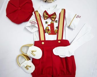 May include: A baby outfit for a first birthday celebration. The outfit includes a white shirt with a gold bow tie, red shorts with suspenders, a red cap, white socks, and gold shoes. The shirt features an embroidered Winnie the Pooh design with the number one and the text "Vishwasha Datta".