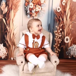 Pode incluir: Um bebê menino vestindo uma roupa marrom e branca com um desenho de leão. Ele está sentado em uma poltrona pequena com uma capa de tecido branco. O fundo é um cenário marrom e bege com uma coroa de flores.