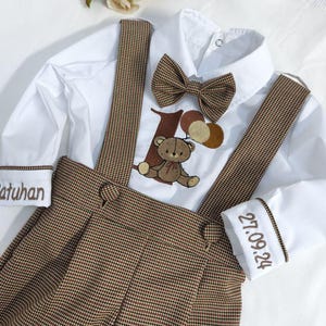 Può includere: Un completo composto da camicia bianca e pantaloncini con bretelle a quadri marroni con papillon abbinato. La camicia presenta un disegno di orsetto e palloncini. I polsini delle maniche sono ricamati con testo e la data 27.09.24.