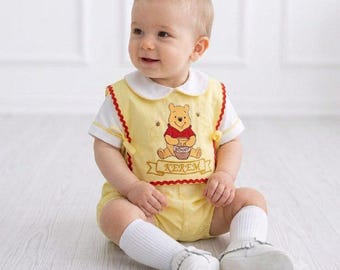 May include: A yellow and white baby romper featuring an embroidered Winnie the Pooh character holding a honey pot. The design includes a red scalloped trim, a white collar, and a banner with the name KEREM embroidered in gold thread on the front.
