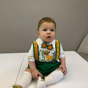 Puede incluir: Un bebé varón con una camisa blanca de manga corta con tirantes verdes y naranjas, pantalones cortos verdes y calcetines blancos. La camisa tiene un número uno verde con un león del Rey León en ella. El bebé lleva una pajarita y zapatos blancos con ribetes dorados.