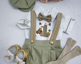 May include: A sage green newsboy cap, white short-sleeve shirt with embroidered Winnie the Pooh, bees, and the number 1, sage green suspender shorts with gold buttons, a matching bow tie, white and gold shoes, and beige socks.