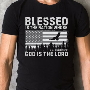 Può includere: Maglietta nera con un design grafico bianco che presenta una bandiera americana usurata, un'aquila che vola sopra e il testo "Blessed is the nation whose God is the Lord" con il versetto biblico "Salmo 33:12" sotto.
