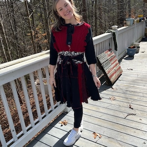 May include: A woman wearing a red and black knitted sweater dress with a black belt. She is standing on a wooden deck with a white railing.