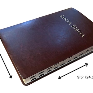 Puede incluir: Una Biblia de cuero marrón con las palabras "SANTA BIBLIA" en letras blancas en la portada. El libro mide 24,5 cm de largo y 18 cm de ancho, con una cinta de marcador.