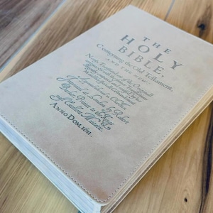 May include: A beige hardcover book titled The Holy Bible, containing the Old and New Testaments. The cover features dark text detailing its 1611 London printing by Robert Barker, printer to the King. The book has stitched edges and a neutral tone.
