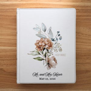 Puede incluir: Una cubierta de libro blanca con un diseño floral con flores melocotón y azules, hojas verdes y las palabras "HOLY BIBLE". La cubierta también incluye los nombres "Mr. and Mrs. Kaiser" y la fecha "MAY 23, 2026."