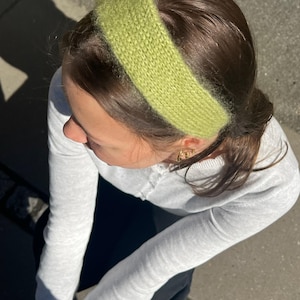 May include: A close-up shot of a person wearing a light green knitted headband. The headband is wide and fluffy, and the person has dark brown hair. The person is wearing a light gray long-sleeved shirt.