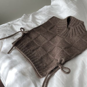 Puede incluir: Un calentador de cuello de punto marrón con un patrón de cuadros. El calentador de cuello tiene lazos a cada lado y está hecho de un material suave y texturizado. Se muestra sobre una sábana blanca.