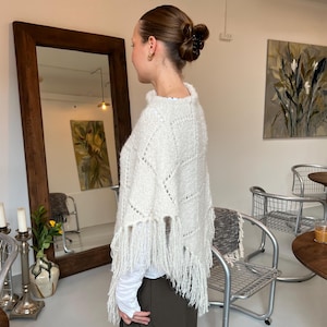 Könnte beinhalten: Ein weißer Strickponcho mit strukturiertem Rautenmuster und langen Fransen am unteren Saum. Das Kleidungsstück wird über einem weißen Langarmshirt und einer dunklen Hose getragen und zeichnet sich durch eine weiche, flauschige Garnstruktur aus.