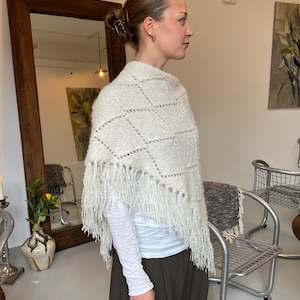 Könnte beinhalten: Ein hellgrauer Strickschal mit Rautenmuster, durchbrochenen Details an den Rändern und langen Fransen. Das Kleidungsstück wird über einem weißen Langarmshirt und einer dunkelbraunen Hose getragen und weist eine strukturierte Strickoptik auf.