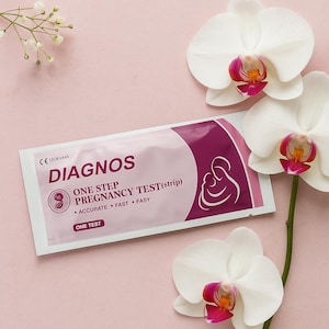 Pode incluir: Um teste de gravidez numa embalagem rosa e branca com a palavra "DIAGNOS" em letras grandes. A embalagem também inclui o texto "ONE STEP PREGNANCY TEST (strip)" e um gráfico de uma mãe e um filho. Orquídeas brancas no fundo.