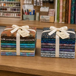 Peut inclure: Deux piles de carrés de tissu pliés attachés avec un ruban crème et une étiquette marquée Batik. La pile de gauche présente des tons chauds, des verts et des bleus. La pile de droite affiche des tons froids de bleu, violet et gris à motifs.