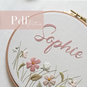 Può includere: Ricamo su telaio con il nome "Sophie" in corsivo rosa, circondato da delicati ricami floreali rosa, verdi e bianchi. Il testo "Pdf Pattern" è in alto a sinistra. Il ricamo è posto in un telaio di legno.