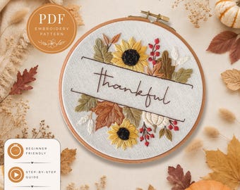 May include: A circular wooden embroidery hoop containing white fabric with a hand-stitched design featuring the word thankful in brown script, surrounded by embroidered yellow sunflowers, green and brown autumn leaves, red berries, and a white pumpkin.