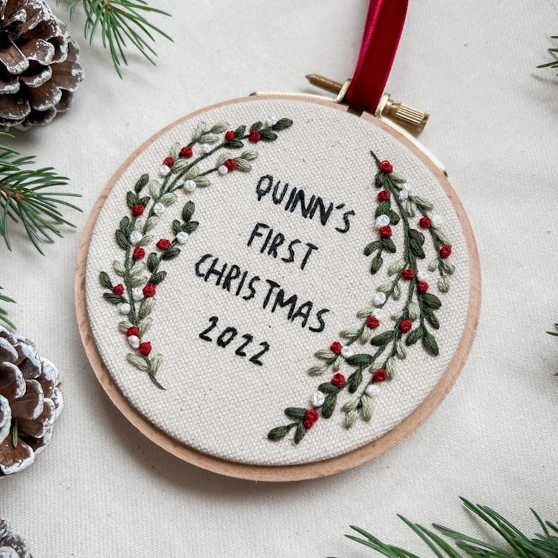 Pu&ograve; includere: Ornamento natalizio ricamato in un cerchio di legno. Il tessuto bianco sporco presenta il testo "Quinn's First Christmas 2022" in nero. Foglie ricamate verdi e bianche con bacche rosse circondano il testo. Un nastro rosso &egrave; attaccato per appenderlo.