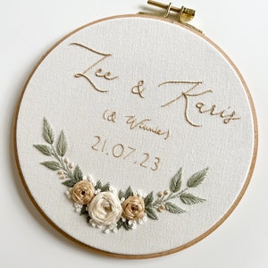May include: Embroidered hoop art with the names "Lee & Kait" and the date "21.07.23" in gold thread. The hoop is decorated with a floral wreath of white and beige roses and green leaves.