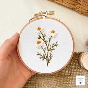 May include: A circular wooden embroidery hoop containing white fabric with a hand-stitched design of three white daisy flowers with yellow centers and green stems. The hoop features a gold-colored metal tightening screw at the top.