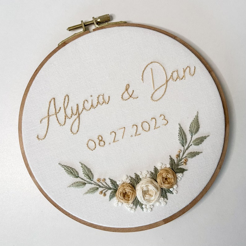 Op de afbeelding: Geborduurde borduurring met de namen "Alycia & Dan" en de datum "08.27.2023" in goudkleurig garen. De witte stof is omlijst door een houten ring en versierd met geborduurde bloemen en bladeren in groen, goud en wit.