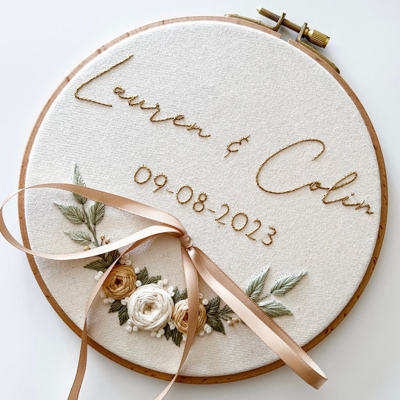 Op de afbeelding: Geborduurde borduurring met de namen "Lauren & Colin" en de datum "09-08-2023" in goudkleurig garen. Het ontwerp bevat een bloemstuk met cr&egrave;me- en bruinroze rozen, groene bladeren en een satijnen strik. De ring is van hout.