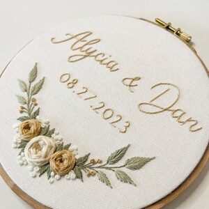 Op de afbeelding: Geborduurde hoepelkunst met de namen "Alycia & Dan" en de datum "08.27.2023" in goudkleurig garen. Het ontwerp bevat een bloemstuk met wit, beige en groen geborduurde bloemen en bladeren, tegen een witte stoffen achtergrond.