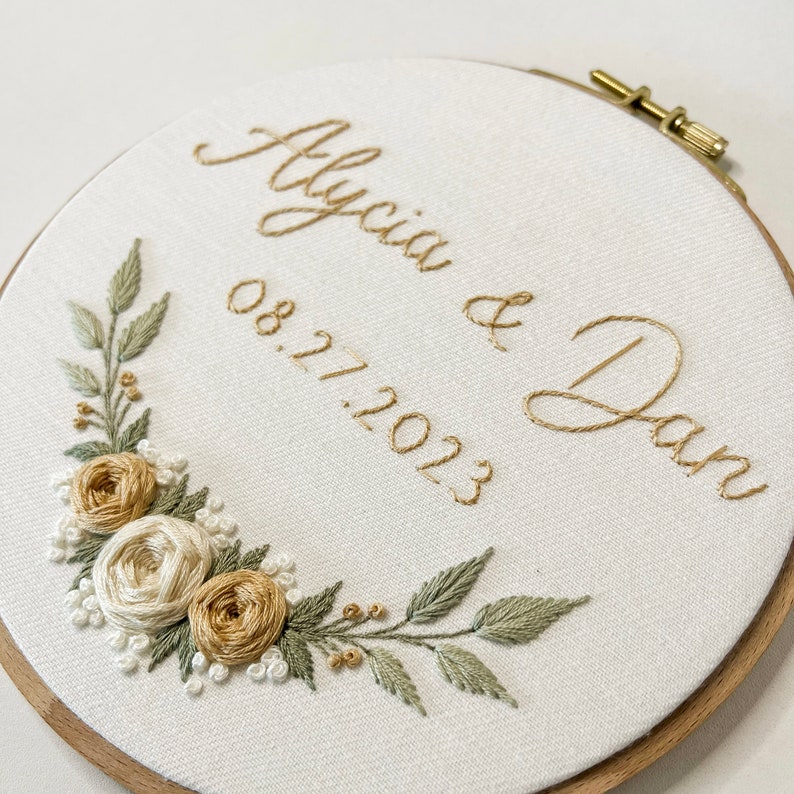 Op de afbeelding: Geborduurde hoepelkunst met de namen "Alycia & Dan" en de datum "08.27.2023" in goudkleurig garen. Het ontwerp bevat een bloemstuk met wit, beige en groen geborduurde bloemen en bladeren, tegen een witte stoffen achtergrond.