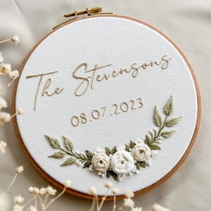 Op de afbeelding: Geborduurde hoepelkunst met de tekst "The Stevensons" en de datum "08.07.2023" in goudkleurig draad. De witte stof is versierd met een bloemen- en bladerontwerp in groen en cr&egrave;me, geplaatst in een houten hoepel.