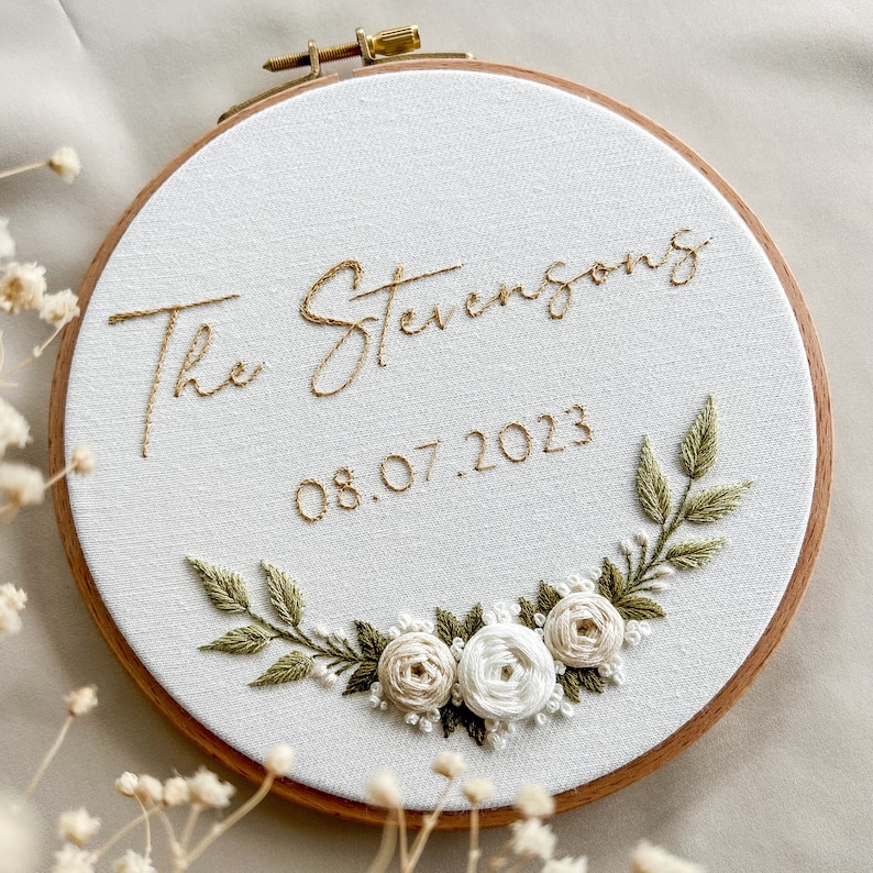 Op de afbeelding: Geborduurde hoepelkunst met de tekst "The Stevensons" en de datum "08.07.2023" in goudkleurig draad. De witte stof is versierd met een bloemen- en bladerontwerp in groen en cr&egrave;me, geplaatst in een houten hoepel.