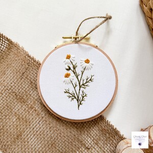 May include: A circular wooden embroidery hoop containing white fabric with a hand-stitched design of three white daisy flowers with yellow centers and green stems. A brown twine loop is attached to the top metal tension screw for hanging.