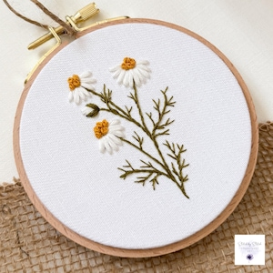 May include: A circular wooden embroidery hoop containing white fabric with a hand-stitched design of three daisy flowers. The flowers feature white petals and textured yellow centers, connected by dark green stems and detailed foliage.