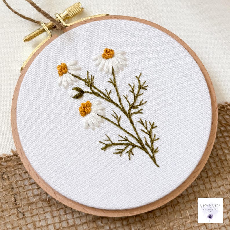 May include: A circular wooden embroidery hoop containing white fabric with a hand-stitched design of three daisy flowers. The flowers feature white petals and textured yellow centers, connected by dark green stems and detailed foliage.