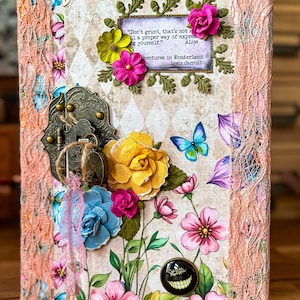 Könnte beinhalten: Ein dekoratives Journal mit Blumen- und Schmetterlingsdesign. Das Cover zeigt ein Zitat aus Alice im Wunderland, einen Metallverschluss und bunte Papierblumen. Die Kanten sind mit pfirsichfarbener Spitze verziert.