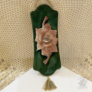 May include: A decorative wall hanging with a large, detailed, peach-coloured rose on a dark green velvet background. The velvet is trimmed with gold rope and has a gold tassel at the bottom. Designed for wall display.