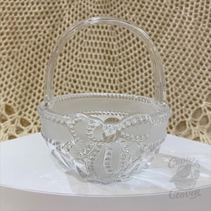 May include: A clear glass basket with a handle, featuring a frosted bow design around the rim. The basket has a textured edge and sits on a white surface. The basket is likely used for decorative purposes.
